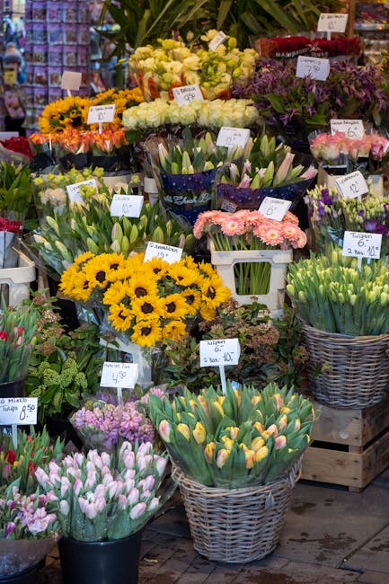A vibrant display of fresh flowers arranged in woven baskets and white plastic containers, featuring yellow sunflowers, pink and white tulips, purple and white hyacinths, and green foliage. The flower
