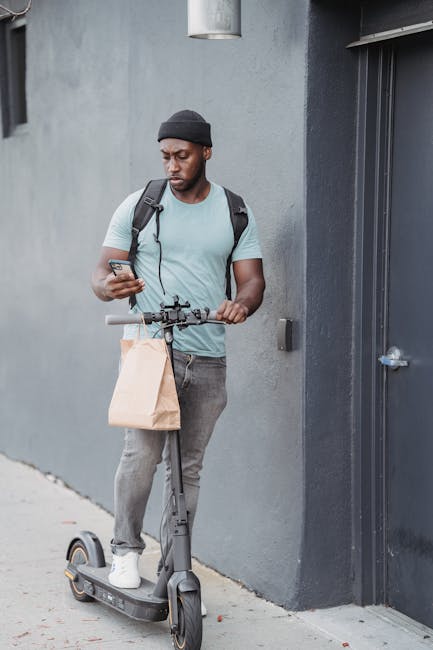 A young man with a dark skin tone, wearing a black beanie, light blue t-shirt, gray jeans, and white sneakers, stands outdoors next to a gray wall and a black door. He is holding a smartphone in his r